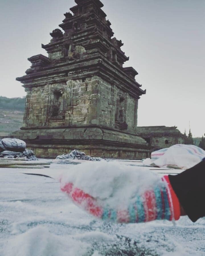 a hand holding snow in front of the Hindu Temple at Dieng Plateau