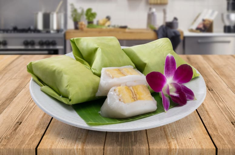 A plate of nagasari - 3 wrapped in a banana leaf and one cut on open. A flower on the side.