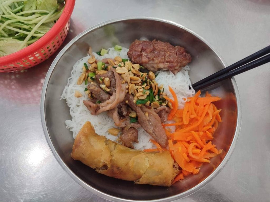 A bowl consisting of a fried spring roll, carrots, noodles, meatball, peanuts, pork meat and herbs.
