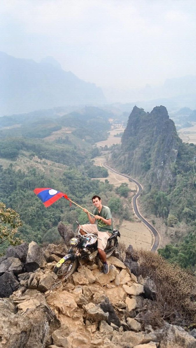 Simon holding the Lao flag on a motorcycle on top of Nam Xay Viewpoint in Laos