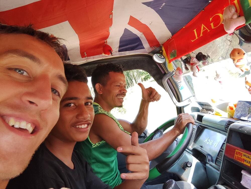 Simon hitchhiking in a van somewhere in Timor Leste. The other 2 men inside show thumbs up and smile.